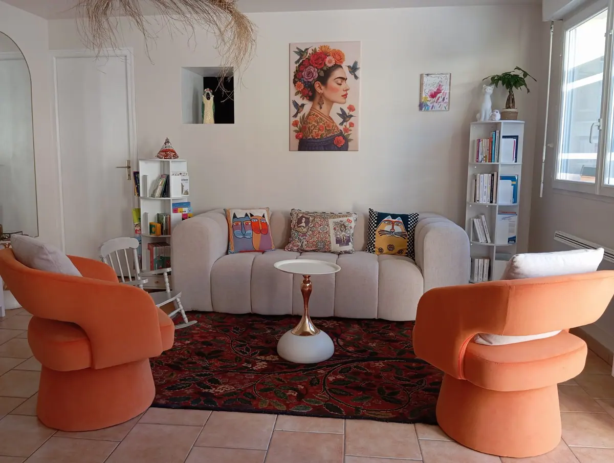 Le salon du cabinet : un canapé clair, deux fauteuils orange et un tapis rouge, avec des étagères de livres et un tableau de fleurs